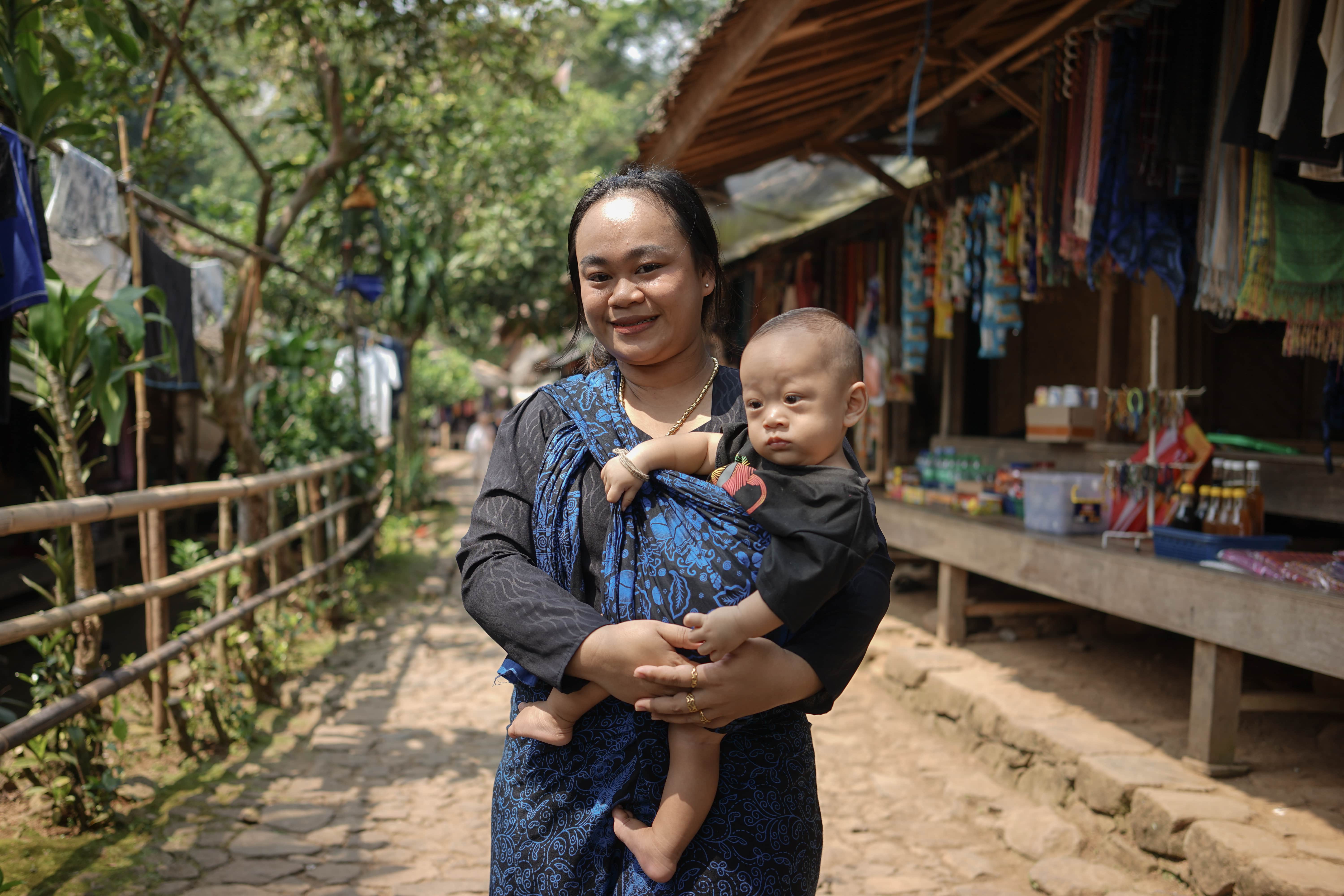 DSC06065-min A woman wearing a black and blue kebaya holding a baby in front of a traditional shop. The background shows a village path with wooden houses, trees, and a peaceful rural atmosphere.